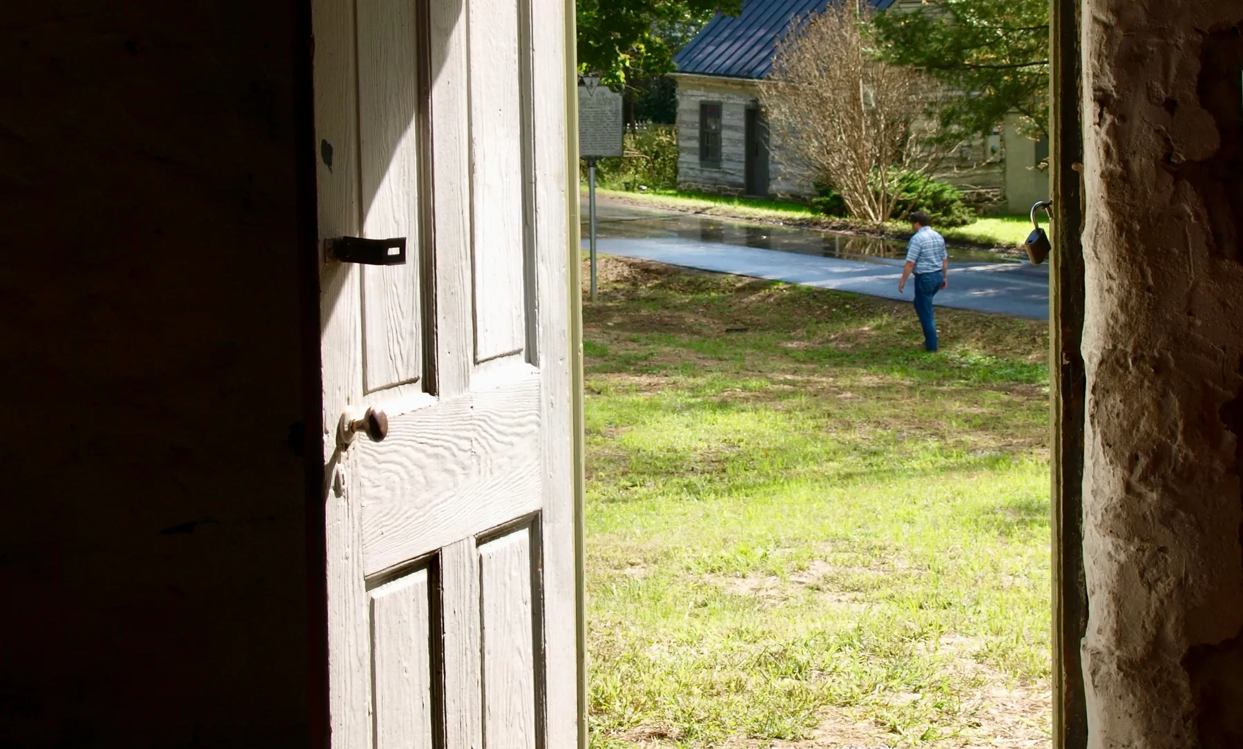 Looking out through an open stone doorway across a sunlit churchyard lawn, with a neighbor walking toward the sidewalk