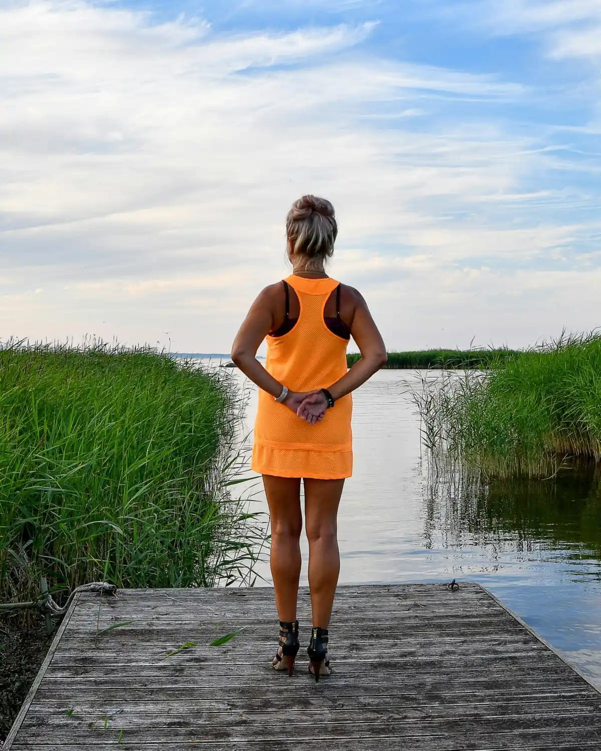 A woman standing at the end of a lake dock at dusk, back to the camera, looking out over the water
