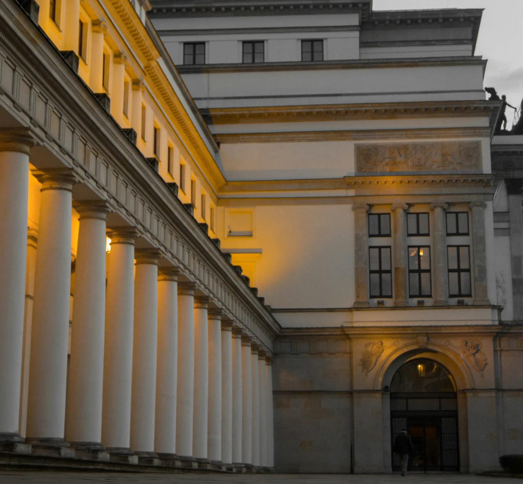 A dramatic low-angle view of a lit colonnade at dusk, with a lone figure walking toward a distant archway entrance.