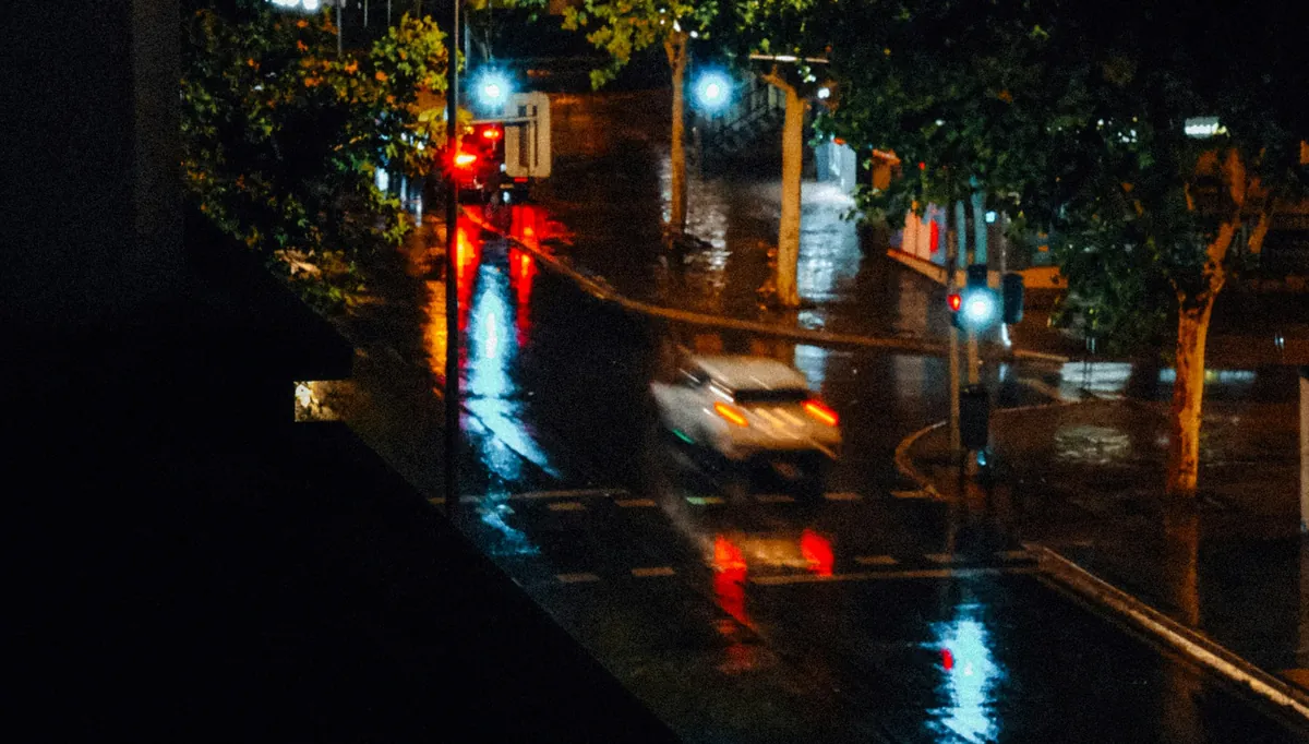 A city street glistening after rain at night, lit by red taillights, streetlamps and a passing car.