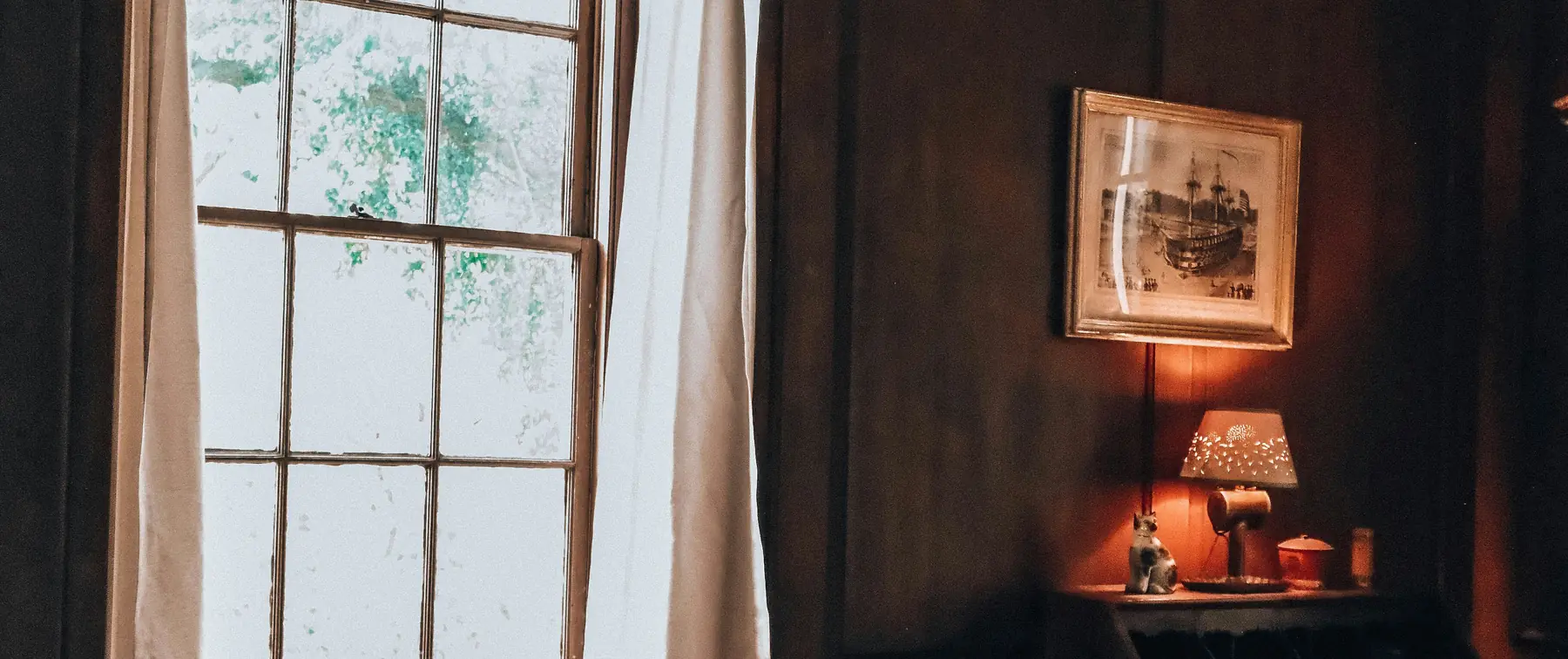 A quiet, wood-paneled reading room with a curtained window on one side and an antique writing desk lit by a small lamp on the other.
